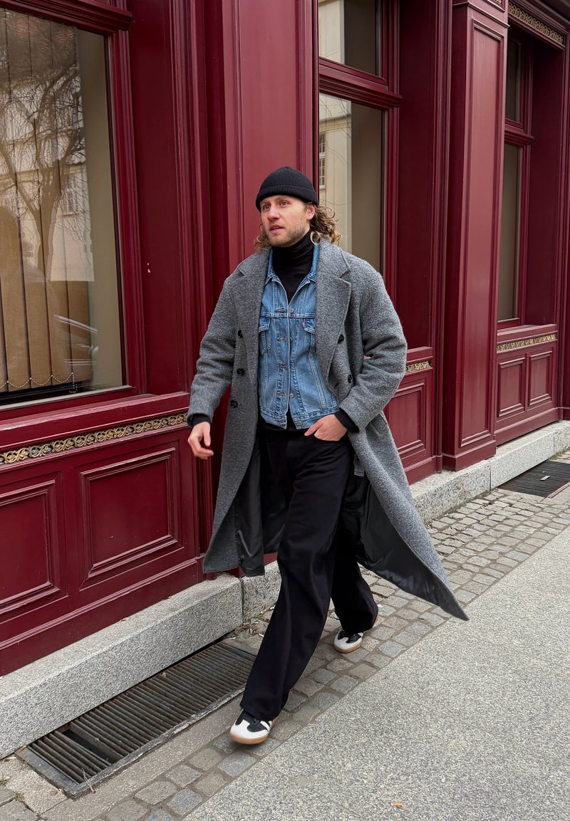 Homme marchant sur un trottoir en ville, portant un long manteau gris, une veste en jean, un pantalon noir, des baskets et un bonnet noir, près de panneaux rouges d'un bâtiment.