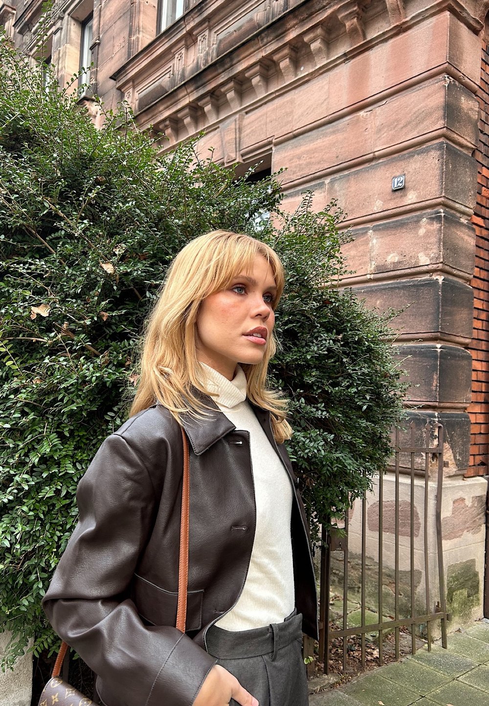 Braune Lederjacke in cropped Form über einem weißen Rollkragenpullover, kombiniert mit grauen Hosen. Der Hintergrund zeigt steinerne Architektur und Grünflächen.