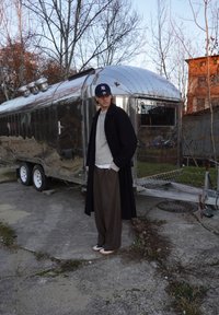 Trailer Airstream argento lucido con dettagli cromati accanto a una persona che indossa un maglione grigio, cappotto nero e pantaloni larghi a strisce marroni.