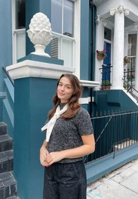 Un top gri texturat și pantaloni negri largi purtați cu un fular alb. În fundal se află o clădire albastră și un obiect decorativ alb în formă de con de pin.