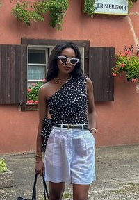 Μαύρη μπλούζα με καφέ κορδόνι και σχέδιο βελόνας στο ένα ώμο, συνδυασμένη με λευκά πλισέ σορτς και μαύρη ζώνη. Τα αξεσουάρ περιλαμβάνουν λευκά γυαλιά ηλίου.