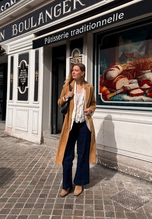 Mujer con abrigo color beige y pantalones azul marino está de pie frente a una panadería francesa con exhibición de pan en la ventana, en una calle empedrada.