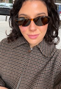 Femme aux cheveux bouclés foncés portant des lunettes de soleil œil de chat écaille de tortue et un manteau marron en pied-de-poule zippé, à l'extérieur sur un trottoir en ville.