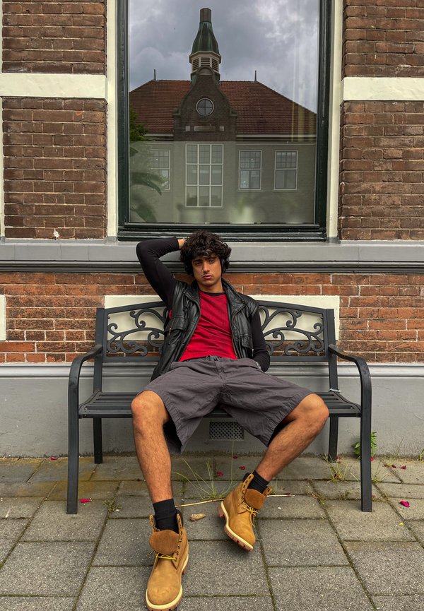Schwarze Metallbank mit kunstvollem Design, eine Person in rotem Shirt und schwarzem Weste sitzt, grauen Shorts und hellbraunen Stiefeln, mit einer Backsteinmauer im Hintergrund.