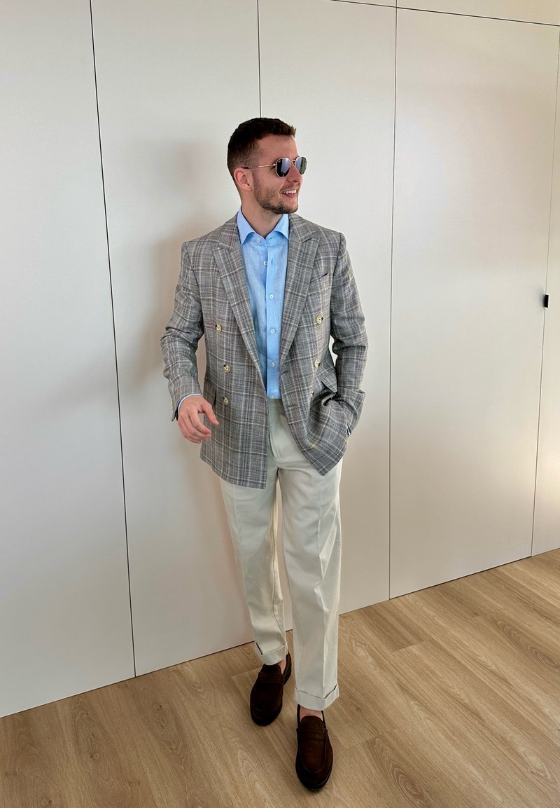 Hombre con gafas de sol, camisa azul claro, blazer cruzado de cuadros, pantalones color crema y mocasines marrones, de pie en el interior sobre suelo de madera.