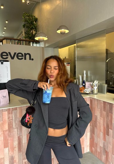 Joven mujer con atuendo oscuro sosteniendo una bebida azul con popote, de pie dentro de una cafetería moderna con un mostrador de azulejos rosas y luces colgantes arriba.