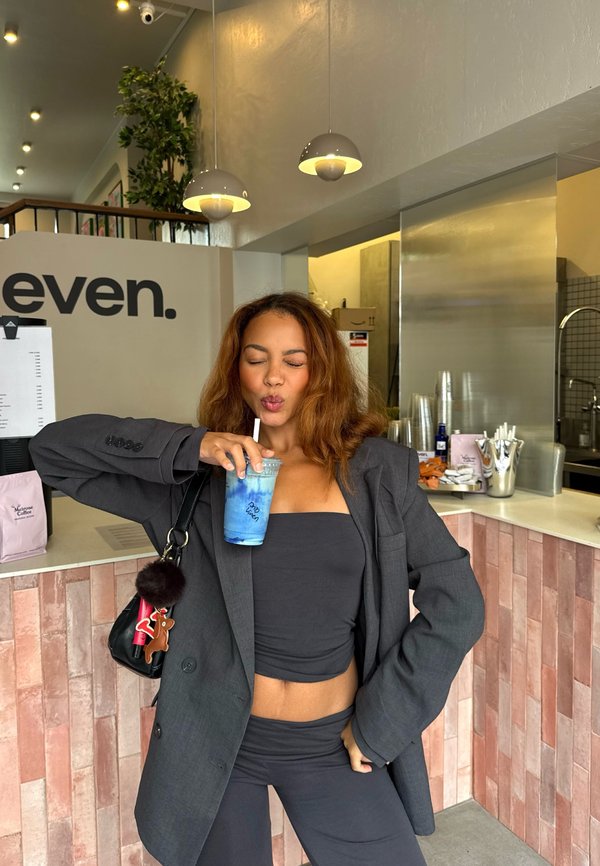 En svart blazer över en crop top, ihop med matchande byxor. Håller en blå drink i en genomskinlig kopp med ett sugrör; modern kafémiljö i bakgrunden.