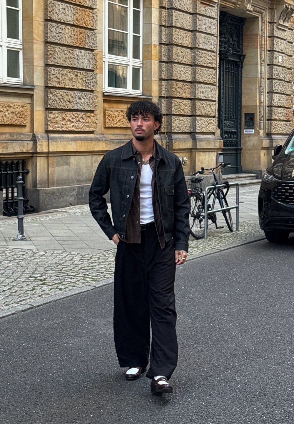 Uomo con i capelli ricci cammina per una strada della città indossando una giacca nera, una maglietta bianca sotto, pantaloni larghi neri e scarpe bicolore marroni e bianche.