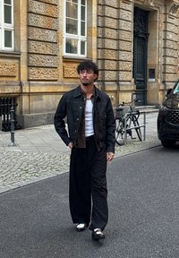 Uomo con i capelli ricci cammina per una strada della città indossando una giacca nera, una maglietta bianca sotto, pantaloni larghi neri e scarpe bicolore marroni e bianche.