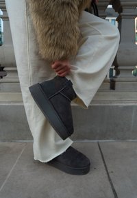 Melni zamšādas potīšu zābaki ar masīvu melnu zolu un potītei paredzētu siksniņu, kombinēti ar krēmkrāsas platām biksēm un teksturētu mākslīgās kažokādas virsdrēbēm.