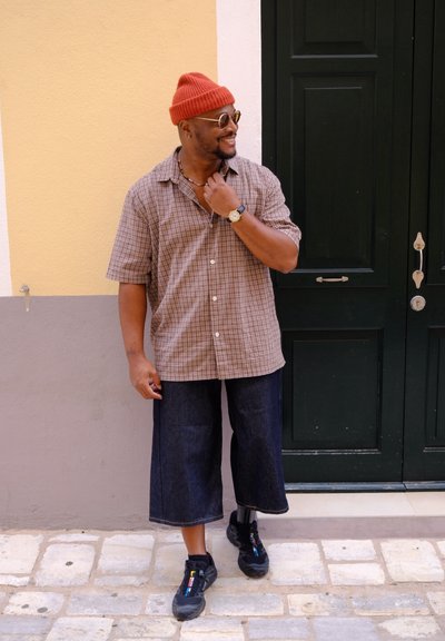 Hombre con gorro rojo, gafas de sol, camisa de cuadros de manga corta, pantalones cortos oscuros anchos y zapatillas, de pie en una calle de adoquines junto a una puerta verde oscura.