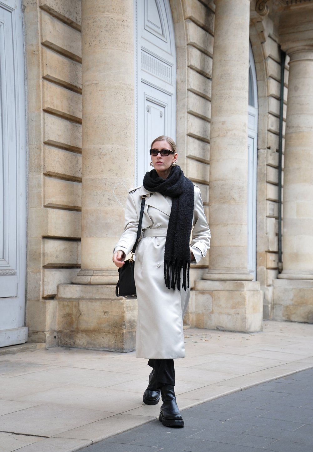 Donna che cammina per una strada cittadina indossando un trench beige, una sciarpa nera, occhiali da sole, stivali neri e portando una borsa nera accanto a colonne di pietra.