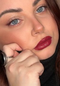 Nærbilde av et ansikt med blå øyne, blanke øyelokk og dype røde lepper. Sølvring på en hånd som hviler på kinnet, kledd i en svart genser med høy hals.