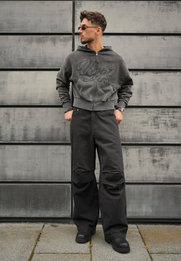 Un homme portant des lunettes de soleil, un sweat à capuche gris zippé, un pantalon noir ample et des chaussures foncées se tient devant un mur en béton.