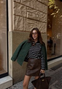 Vrouw met een bril, gestreepte shirt, groene jas en een beige leren rok, die een donkerbruine handtas vasthoudt buiten een stenen gebouw met grote ramen.