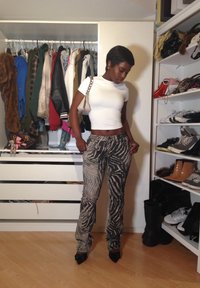 Weißes kurzärmeliges Cropped-Top, gemusterte grau-schwarze Zebra-Streifen-Hosen, schwarze spitze Absätze, Taschen und Kleiderständer im Hintergrund.