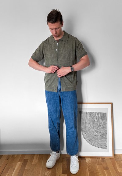 Hombre con camisa verde de manga corta y vaqueros azules abotonándose la camisa, de pie sobre un suelo de madera cerca de una obra de arte abstracta en blanco y negro enmarcada.