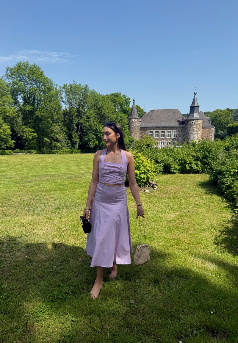 Femme en robe lavande marchant pieds nus sur l’herbe, tenant deux sacs à main, avec un château en pierre et des arbres verts en arrière-plan sous un ciel bleu clair.