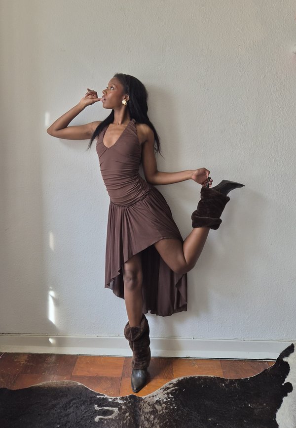Woman in brown dress and fur boots poses on one leg against white wall, holding her raised foot with one hand and touching her mouth with the other.