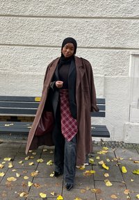 Lungo cappotto in suede marrone indossato sopra un top nero, con una sciarpa a scacchi rossa e nera tessuta, pantaloni a gamba larga neri e décolleté a punta.