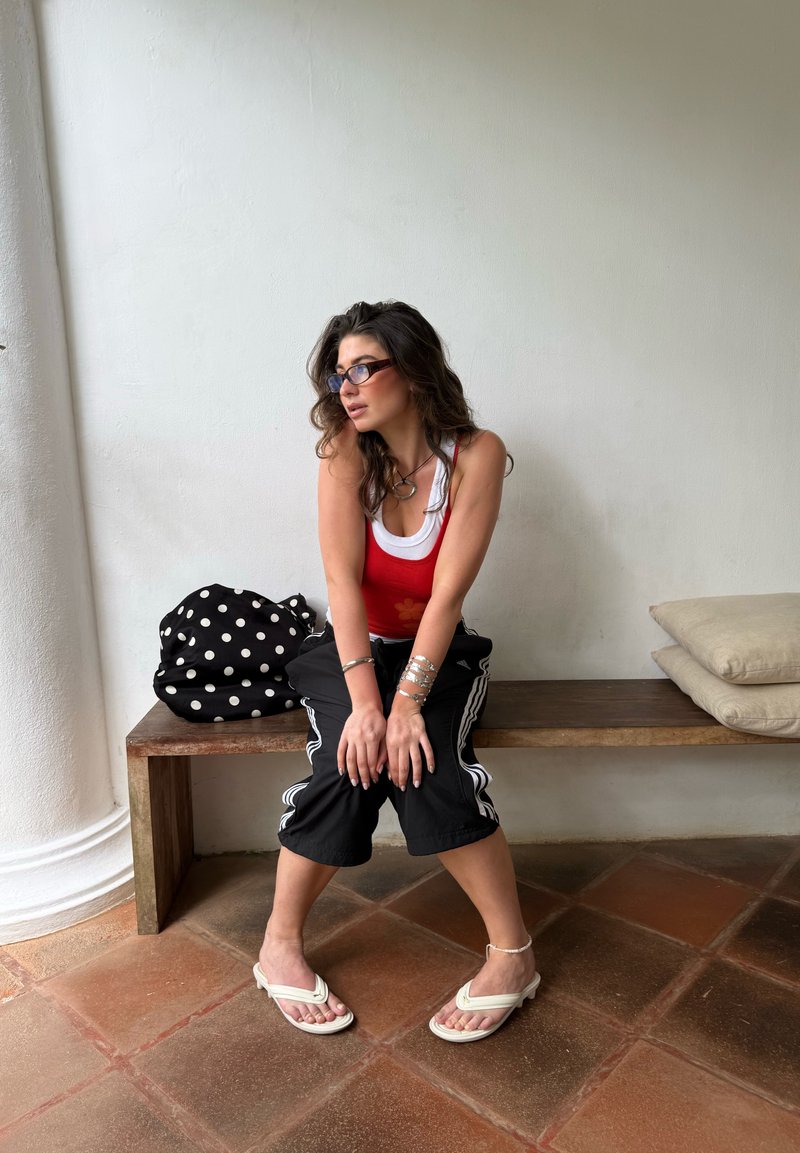 Jeune femme aux longs cheveux bruns portant des lunettes, un débardeur rouge, un pantalon noir et des sandales blanches, assise sur un banc en bois avec un sac à pois et des coussins.