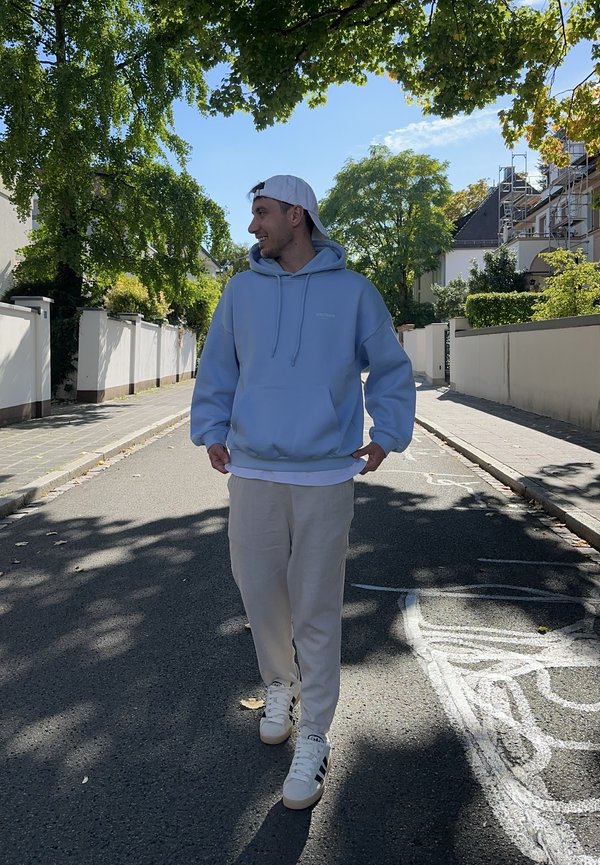 Light blue hoodie with drawstrings, white undershirt, beige trousers, and white trainers. Standing on a sunlit street with trees.