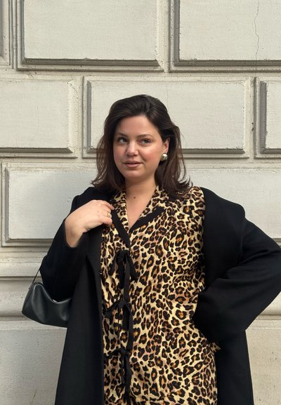Mujer de pie contra una pared beige, vistiendo una blusa de estampado de leopardo, abrigo negro, pendientes de oro y llevando un bolso negro de hombro.