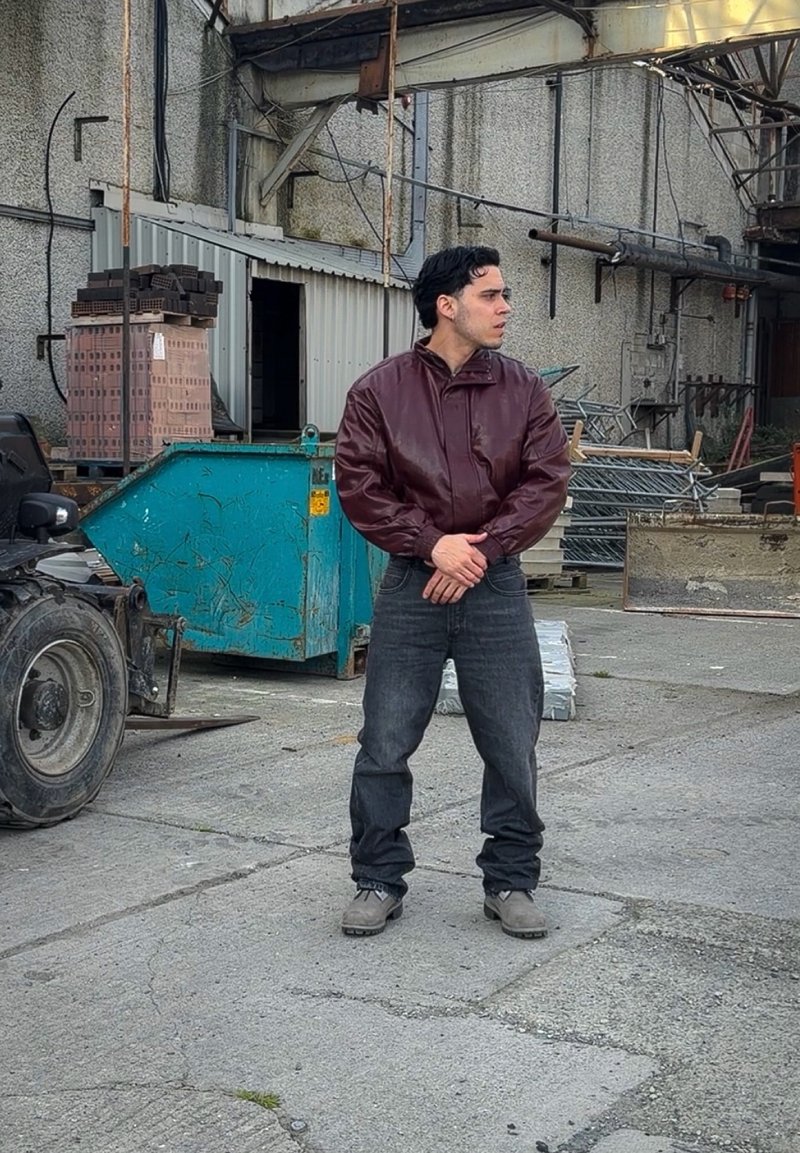 Man draagt een bordeauxrode jas en donkere jeans en staat in een industriële opslagplaats nabij pallets, metalen voorwerpen en een blauwe container.
