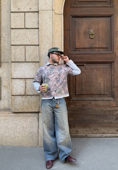 Hombre con gorra, gafas de sol, camisa estampada, vaqueros anchos y zapatos marrones sostiene una bebida, parado junto a una gran puerta de madera con aldaba en forma de cabeza de león.