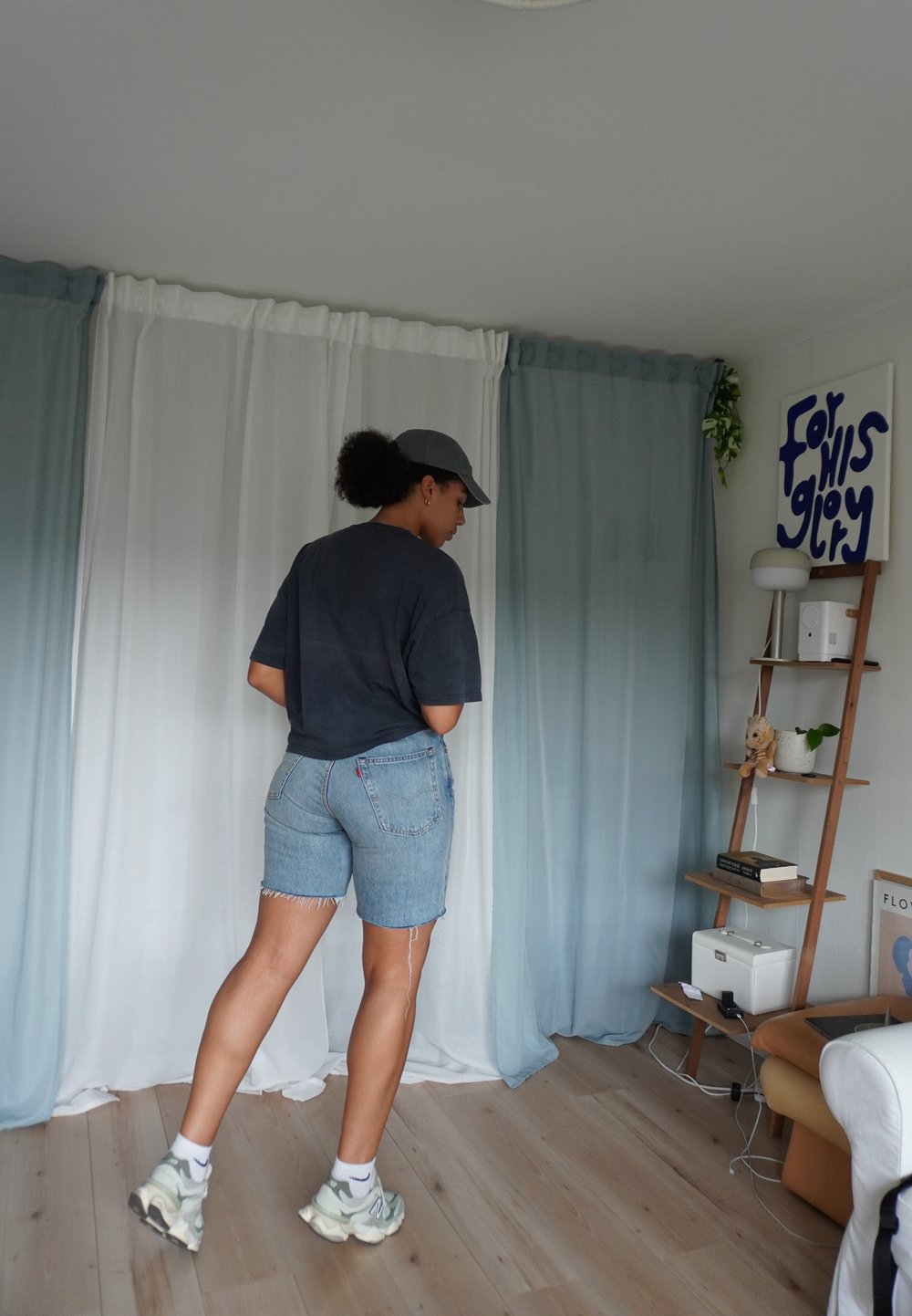Tricou supradimensionat de culoare gri închis, pantaloni scurți din denim albastru cu margini desfăcute, adidași albi și gri. Fundal cu perdea moale și raft din lemn.