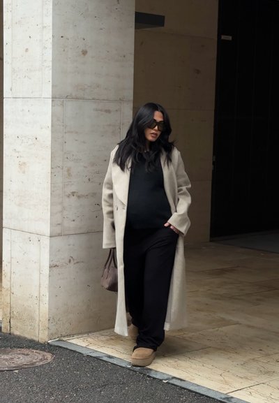 Mujer con abrigo beige, conjunto negro y gafas de sol caminando cerca de la entrada de un edificio con paredes de piedra y suelo de baldosas.
