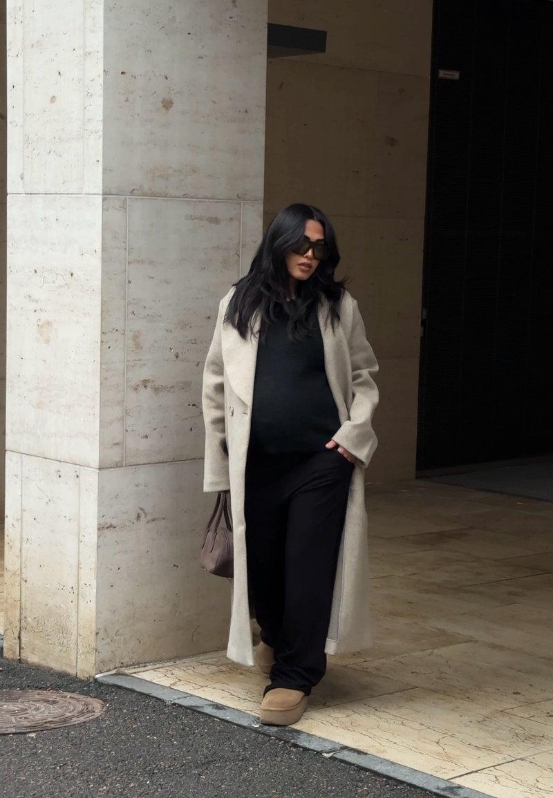 Femme en manteau beige, tenue noire et lunettes de soleil marchant près d'une entrée de bâtiment aux murs en pierre et au sol carrelé.