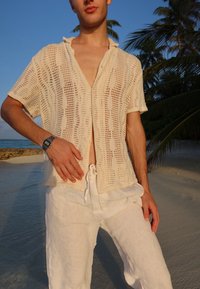 Cream-coloured crochet shirt with a loose fit and short sleeves, paired with white linen trousers. Visible texture and open-knit design.