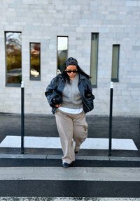 Czarna skórzana kurtka na szarym swetrze, beżowe spodnie o szerokich nogawkach i czarne buty. Wzorzysty chustka na głowie i trzyma czarną torebkę.