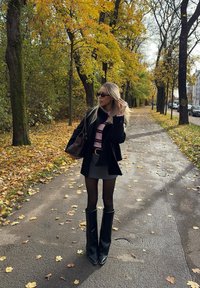 Črni plašč, roza pulover z črtami, siva krilo, črne nogavice, črni škornji do kolen. Rjava torbica. Pot obložena z jesenskim listjem.