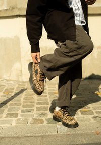 Schuhe mit Leopardenmuster, schwarzer Sohle und Schnürsenkeln, kombiniert mit grünen Cargohosen. Das Design hat eine strukturierte Oberfläche und eine abgerundete Form.