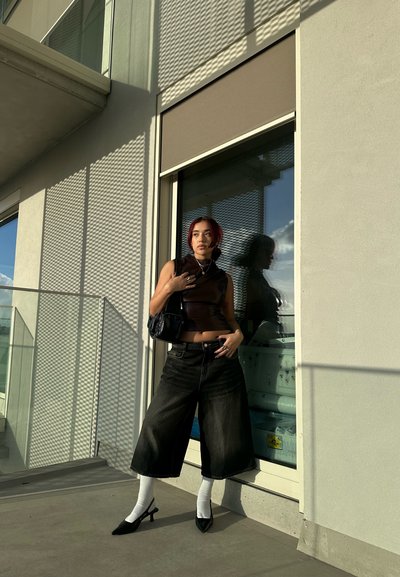 Jeans de pierna ancha cortos de color negro, top sin mangas oscuro, calcetines tobilleros blancos y tacones de punta negra. Pequeño bolso negro; ventana de cristal de fondo.