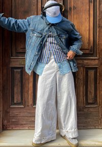 Casaco de denim, camisa riscada e calças largas brancas. Boné azul com riscas claras. Footwear cinza e auscultadores que adicionam contraste. Fundo em madeira.