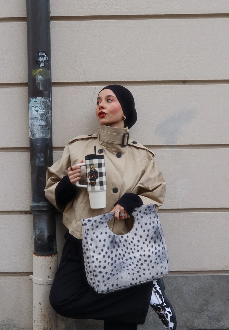 Femme portant une veste beige et un foulard noir, tenant une tasse de voyage à carreaux et un sac à main gris tacheté, debout contre un mur beige.