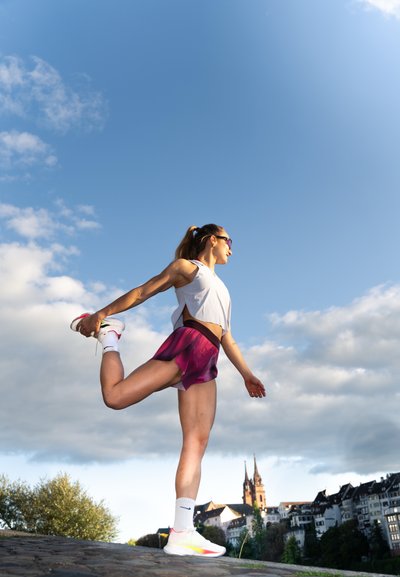 Atleta estirándose en un camino de piedra, vistiendo una camiseta blanca corta y pantalones cortos rosas. Zapatillas blancas con acentos amarillos y calcetines hasta la rodilla.