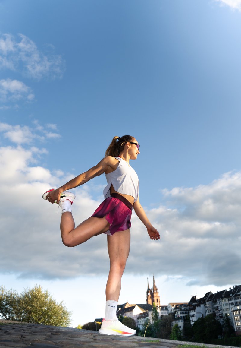 Atlet stretching na kamenné cestě, oblečený v bílém zkráceném topu a růžových šortkách. Bílé tenisky se žlutými akcenty a ponožky až po kolena.