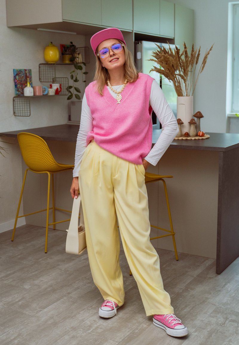 Oversized rosa gestrickte Weste über einem weißen Langarm-Shirt, gelbe plissierte Hose, rosa Mütze, runde Brille und rosa Sneaker.