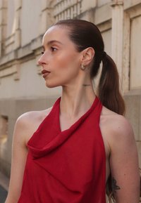 Top rosso senza maniche con tessuto drappeggiato. Il modello ha una coda di cavallo, orecchini discreti e tatuaggi sul collo e sul braccio, mostrato di profilo.