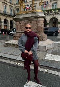 Szary, oversize sweter, bordowa szal, plisowana szara spódnica, burgundowe rajstopy i lakierowane szpilki, trzymająca bordową torebkę z pikowaniem.