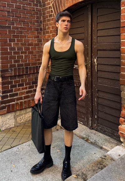Shorts de mezclilla negra, top de tirantes oliva, cinturón negro, bolsa tote negra. Zapatillas negras elegantes y calcetas altas completan el look frente a un fondo de ladrillo.