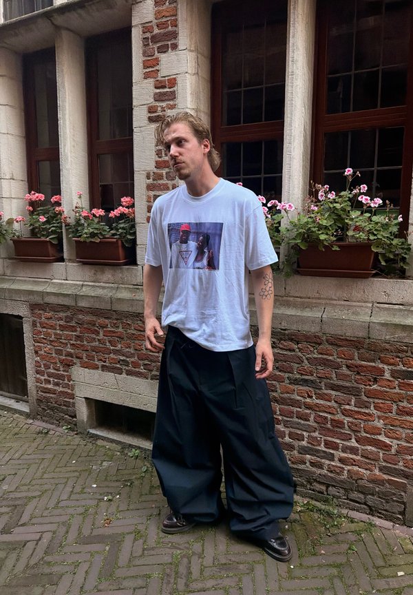Ung mand med lyst hår står på brostensbelægning iført en hvid T-shirt med print, oversize sorte bukser og sorte sko ved blomsterpyntede vinduer.
