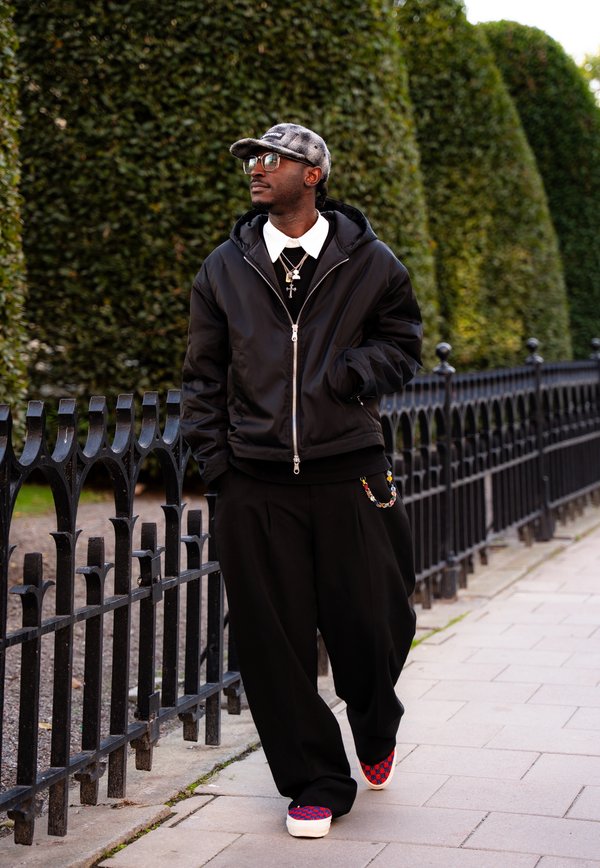 Homme portant une veste noire, une chemise blanche à col, un pantalon foncé et une casquette grise marchant près d'une clôture métallique noire et de hauts buissons verts taillés.