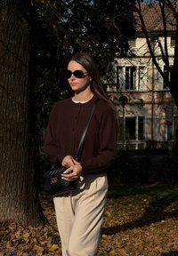 Brun strikket cardigan, hvid skjorte, cremefarvede bukser, sort quiltet håndtaske, runde sorte solbriller, stående ved siden af et træ i et udendørs miljø.