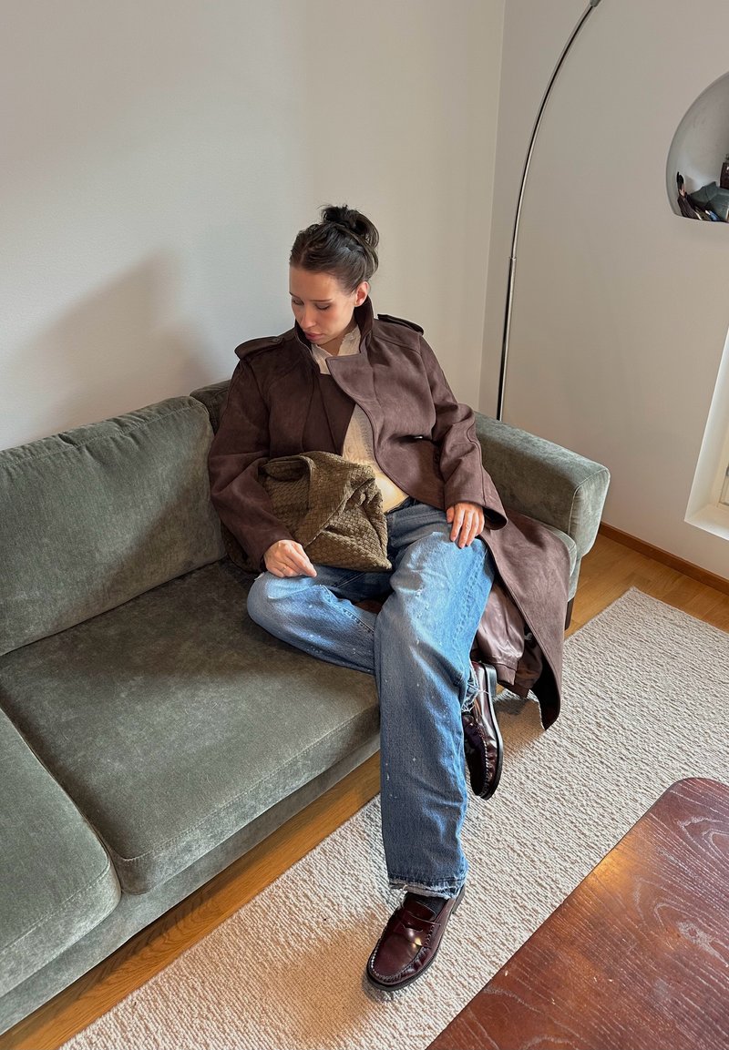 Manteau en suède marron avec épaulettes, pull léger, jean bleu usé et chaussures foncées, assis sur un canapé en velours vert.