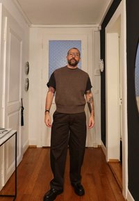 Brązowy sweter na ramiączkach z czarną podkoszulką, zestawiony z brązowymi spodniami i czarnymi butami. Widoczna drewniana podłoga i białe ściany.